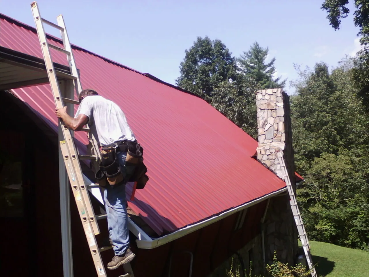 Roofer on a ladder beside a red metal roof and stone chimney — Soffit & Fascia Repair by Roofing Brothers in Enola
